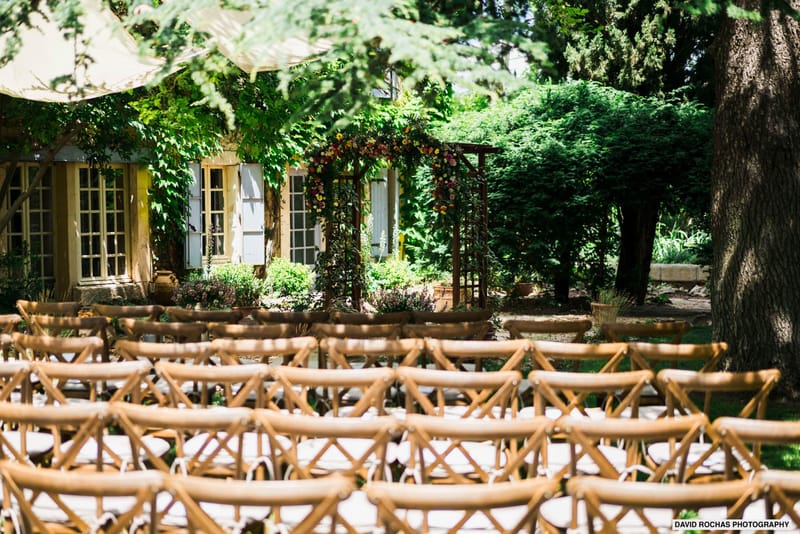 An outdoor wedding ceremony setup photographed before guests arrive, set in a garden in front of a French country-style stone building with pale blue shutters and ivy-covered walls. Multiple rows of natural wood cross-back chairs are arranged facing a wooden arch decorated with a vibrant floral arrangement featuring deep pink, red, orange, and yellow blooms. The wide shot is taken from behind the seating area looking toward the floral arch and building, with the foreground chairs slightly out of focus to draw the eye toward the ceremony backdrop. The overall styling is rustic-garden with warm natural wood tones and a colorful, loose floral palette. Potential venue feature image.