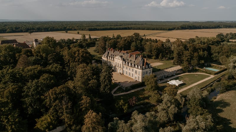 An aerial drone shot capturing a French château and its surrounding grounds in full. The château is a multi-storey classical French manor with a slate mansard roof, terracotta chimney stacks, ornate stone façade detailing, and tall arched windows with shutters. A gravel forecourt sits at the front entrance, and a long lower wing extends to the right side of the main building. On the right side of the grounds, a rectangular swimming pool is visible, along with a white marquee or stretch tent set up on the lawn, suggesting wedding event use. The property is surrounded by mature trees on multiple sides, with open agricultural fields visible in the background. Potential venue feature image.