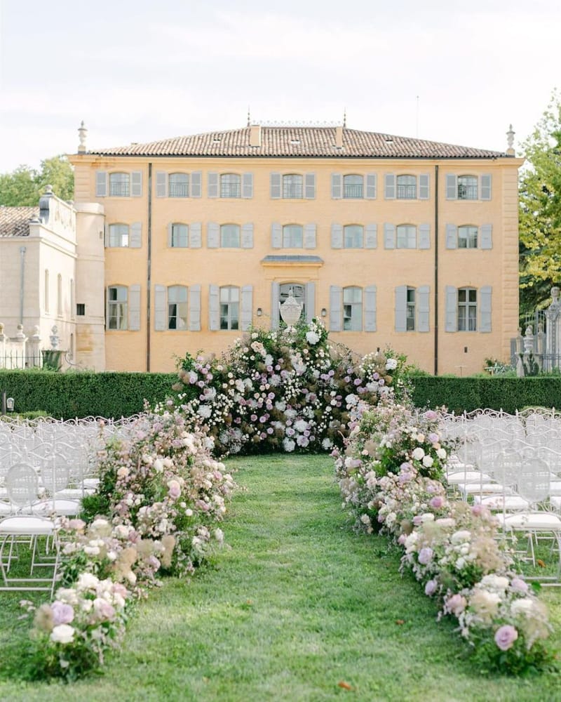An outdoor wedding ceremony setup on a formal lawn in front of a three-story French château with warm ochre-yellow render and pale blue-grey shutters. The aisle is flanked by two curved floral arches and low ground-level floral runners, all composed of blush pink roses, mauve and lilac roses, ivory white blooms, pampas grass, and trailing greenery. At the altar, a large dense floral installation surrounds a white stone urn pedestal, continuing the same blush, mauve, and ivory palette with abundant foliage. White ghost-style acrylic chairs with white cushions are arranged in rows on either side of the aisle, accommodating what appears to be 60–80 guests. The overall styling is romantic and garden-inspired with a soft pastel color palette. Wide establishing shot taken from the entrance of the aisle looking toward the altar and château facade. Potential venue feature image.