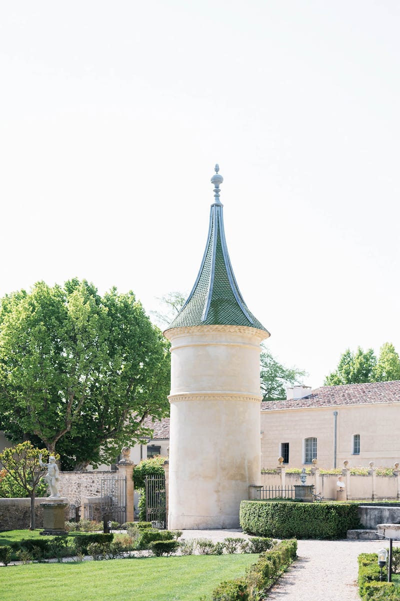 French chateau stone turret with pointed green roof flanked by formal boxwood gardens and gravel path