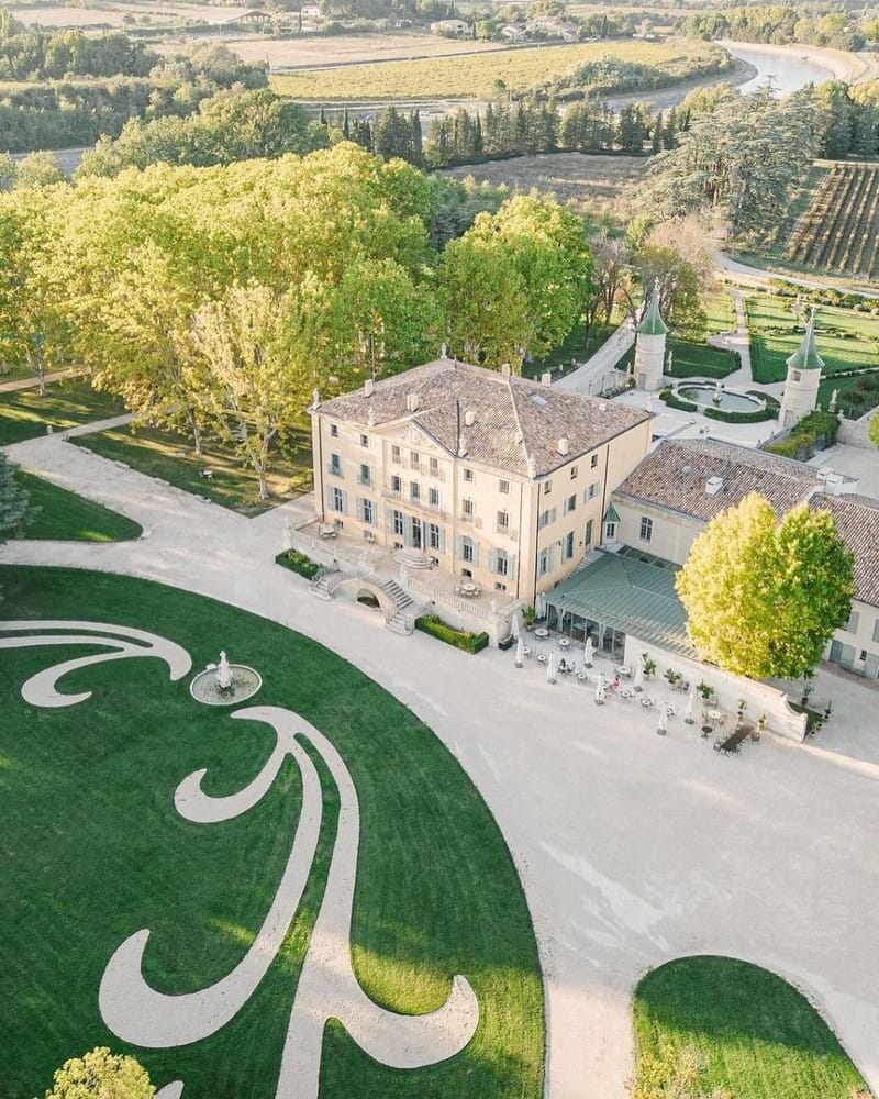 Aerial view of pale yellow chateau with parterre garden, fountain, and green-roofed turrets amid vineyards