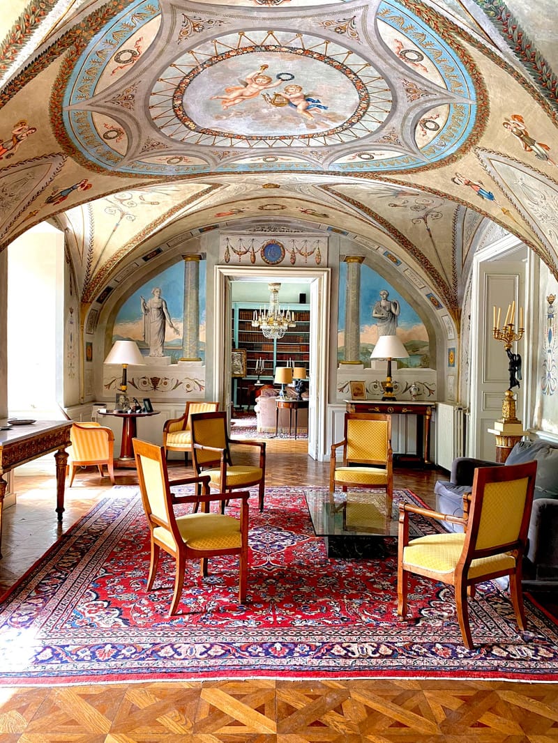 An interior room of a historic château or grand manor house, photographed with a wide-angle vertical composition showing no people or wedding activity. The room features a vaulted ceiling covered in elaborate neoclassical painted frescoes depicting cherubs, classical figures, garlands, and geometric patterns in sky blue, gold, terracotta, and cream tones. The walls carry painted murals of classical female figures set between painted columns. The furnishings include mahogany Empire-style armchairs upholstered in mustard yellow fabric, arranged around a low glass-topped coffee table atop a large red and navy Persian-style rug. A gilded console table with a white lamp sits against the far wall, flanked by matching lamps. Parquet flooring with inlaid geometric marquetry is visible in the foreground. Through a central doorway, a book-lined library with a crystal chandelier is visible. The overall interior style is French neoclassical. Potential venue feature image.
