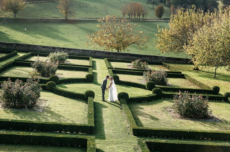 Elevated view of bride and groom kissing in geometric boxwood parterre garden with autumn trees