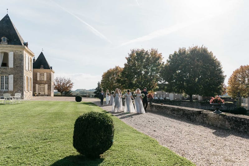 Bride and powder blue bridesmaids walking gravel path toward slate-roofed chateau with topiary gardens