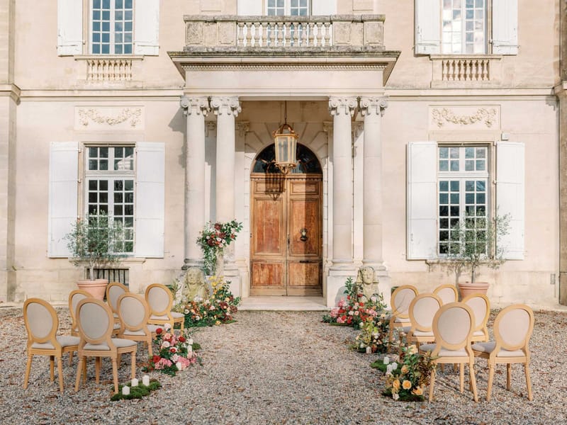 Ceremony setup with burgundy and coral ground florals, oak Louis XVI chairs, and olive trees at chateau door