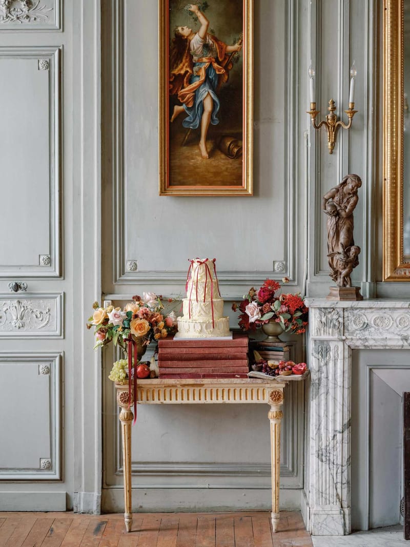 Three-tiered ivory wedding cake with red ribbon on gilded console beside burgundy and peach floral arrangements