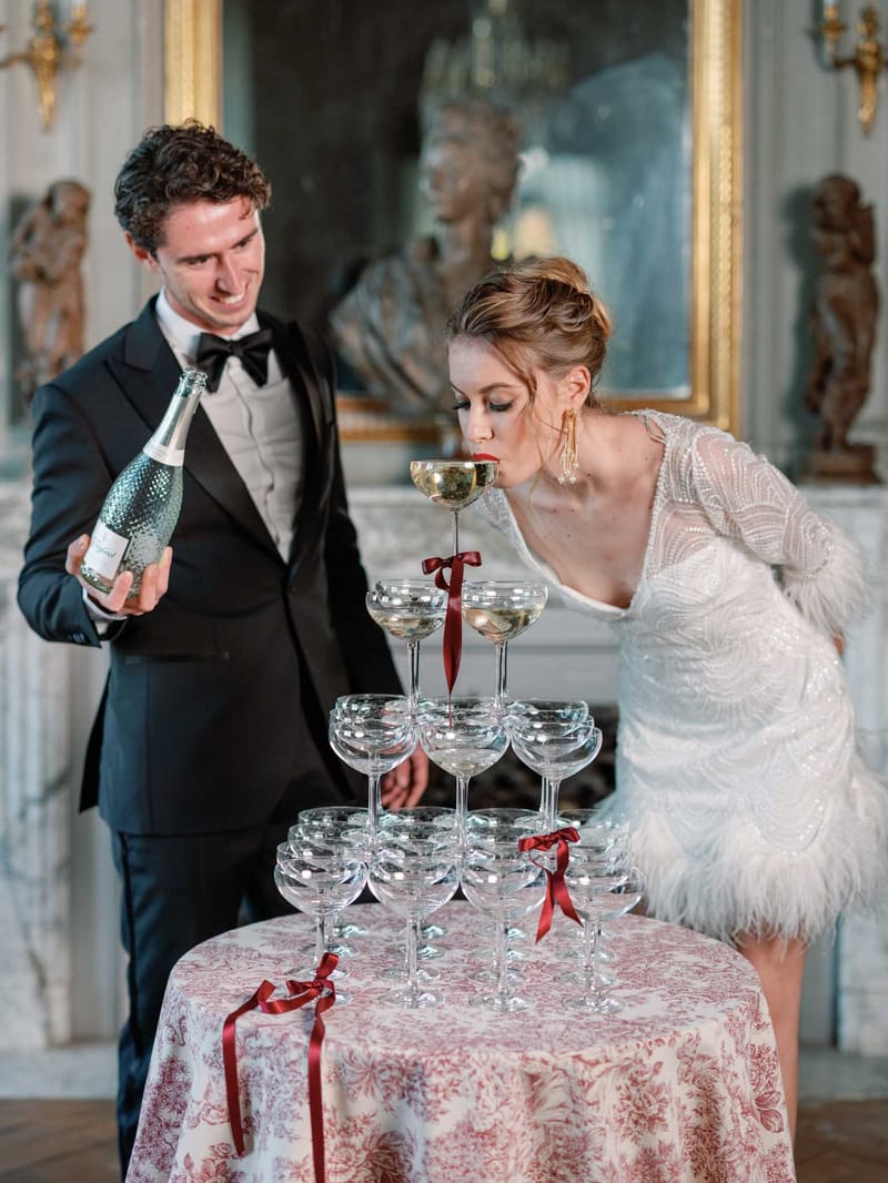 Bride sipping from champagne tower while groom pours in ornate gilded French chateau interior