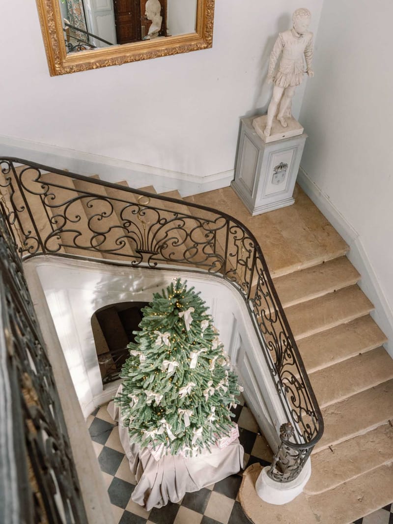 An overhead wide shot of a château interior staircase hall, captured from an upper floor looking down. The focal point is a large Christmas tree decorated with warm fairy lights and ivory ribbon bows, wrapped at the base with a linen-toned skirt, positioned at the foot of a curved stone staircase with an ornate wrought-iron balustrade featuring scrollwork detailing. The floor below is laid in a black-and-white diamond-pattern stone tile. A white marble statue of a classical figure stands on a pale grey carved pedestal on the landing. A large gold-framed mirror with carved detailing hangs on the white wall above. No people are present in the image. Potential venue feature image.