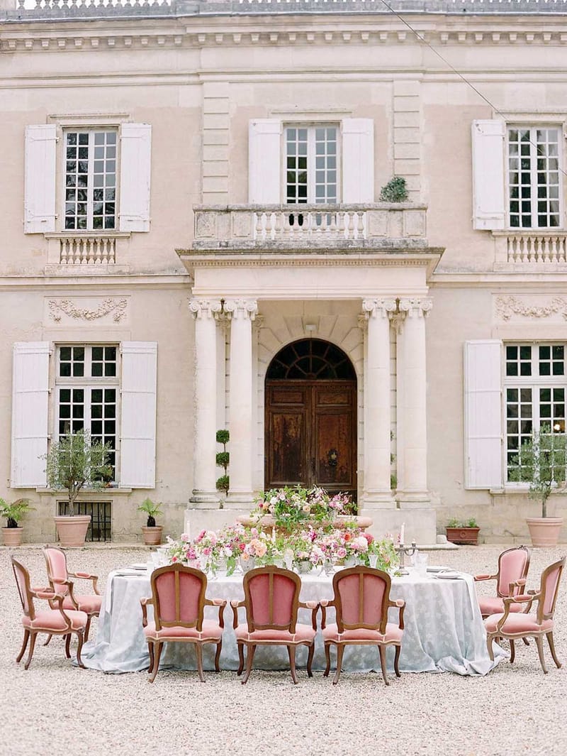 Oval dining table with dusty rose velvet chairs and coral rose centerpiece before a classical French chateau entrance