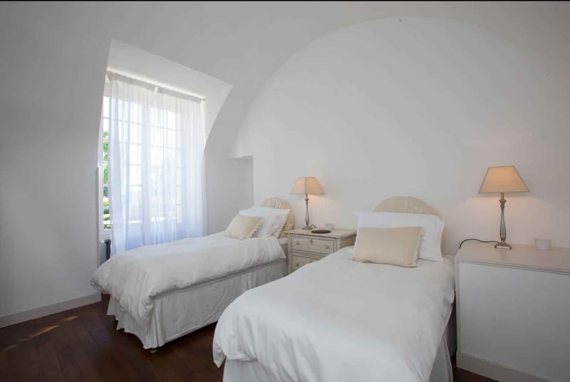 Interior wide shot of a twin bedroom inside what appears to be a French château or manor house, featuring a vaulted barrel ceiling finished in white plaster. The room contains two single beds dressed in white linen with cream ruffled accent pillows and cream upholstered headboards, flanked by a painted grey-blue nightstand and a white bedside table, each topped with a warm-toned beige table lamp. A tall arched window with white sheer curtains allows natural daylight into the space, and dark hardwood flooring contrasts with the all-white walls and ceiling. The décor palette is white, cream, and soft grey with a classic French country styling. Potential venue feature image.