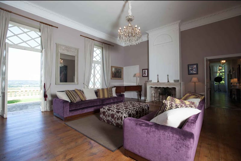 Interior wide shot of a formal salon inside what appears to be a French château, showing a high-ceilinged reception room with pale lavender-grey walls, white ornate crown molding, and wide-plank hardwood floors. The room is furnished with purple velvet sofas and armchairs, gold and dark patterned scatter cushions, and a floral-upholstered ottoman in white and aubergine. A crystal chandelier hangs from the ceiling, and a carved stone fireplace with candle holders sits along one wall, flanked by framed paintings and a gilt-framed mirror. Full-height diamond-pane windows and open French doors lead to a balustrade terrace with a panoramic countryside view beyond. Potential venue feature image.