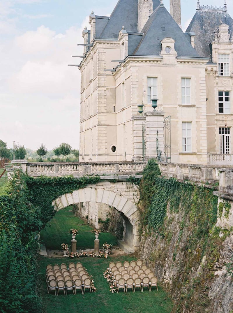 Ceremony setup in chateau courtyard with Chiavari chairs facing stone archway bridge and floral urns, cream facade behind