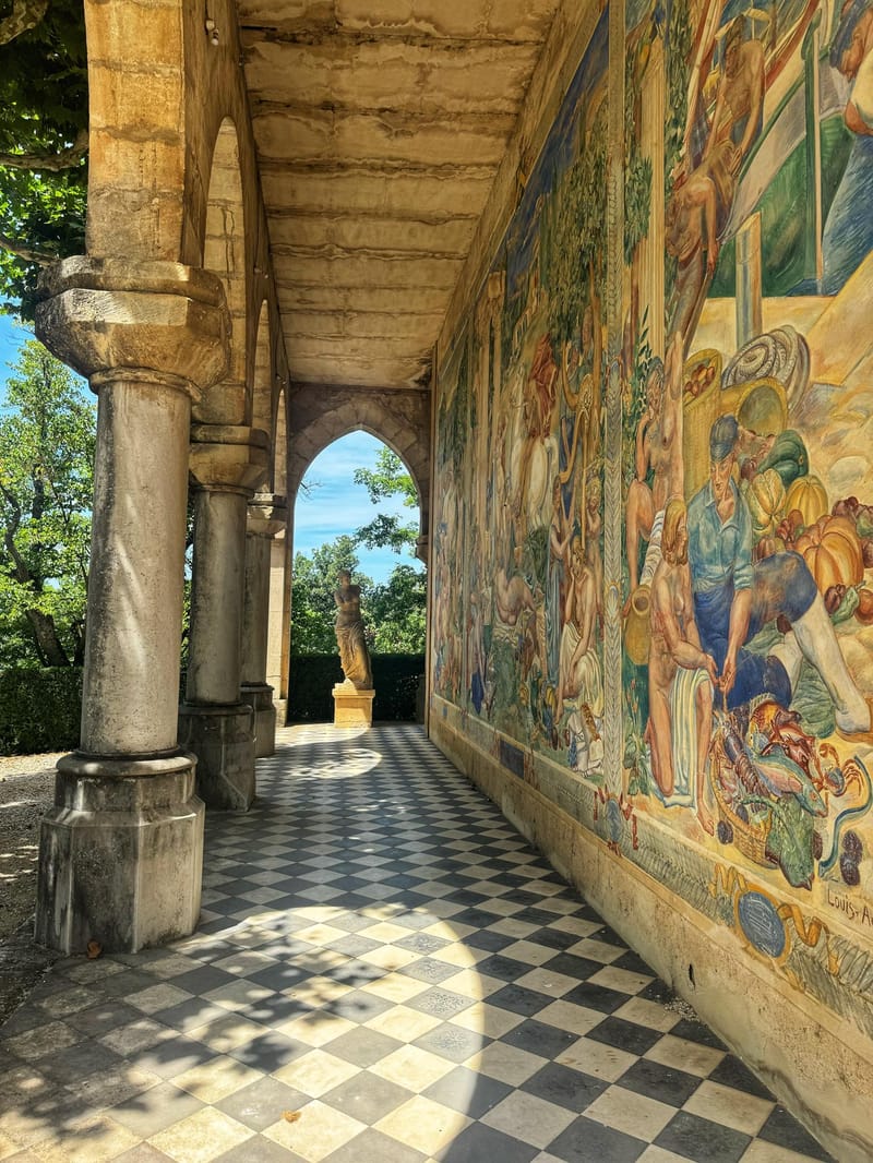 A wide shot of an outdoor covered gallery or loggia at a historic French venue, featuring no people or wedding activity. The architectural space is defined by a black-and-white checkered tile floor, stone columns with carved capitals, and a vaulted stone ceiling. The right-hand wall is covered in a large-scale painted mural depicting figures with produce, seafood, and harvest scenes in warm ochre, green, and blue tones, signed 'Louis-A' at the lower right. At the far end of the colonnade, a pointed Gothic arch frames a classical female statue on a pedestal, with lush green garden foliage and blue sky visible beyond. The composition is a perspective shot leading the eye down the length of the gallery. Potential venue feature image.
