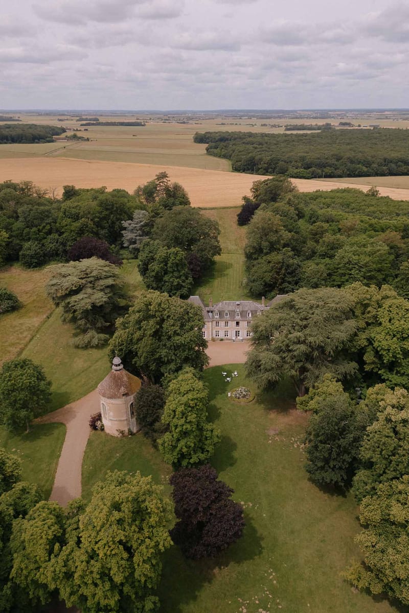 Aerial view of French chateau estate with rose-toned manor house surrounded by mature parkland and farmland