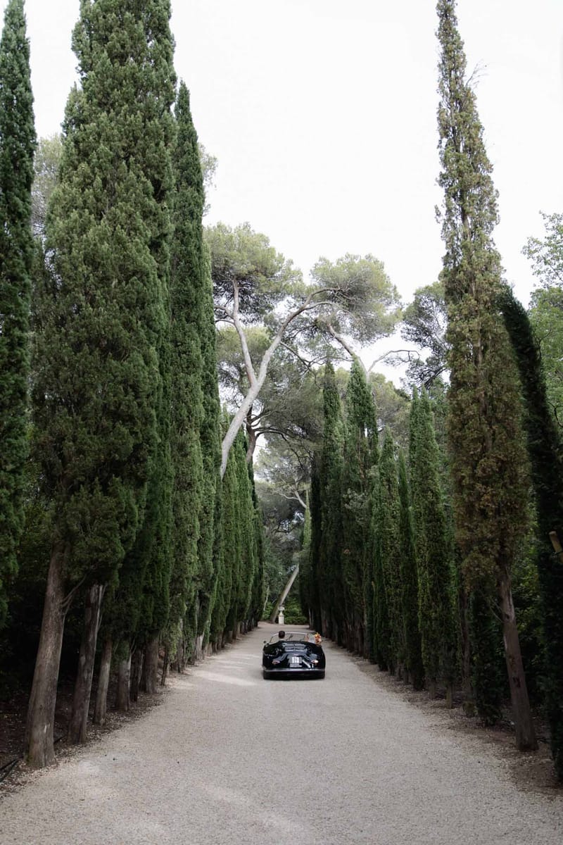 Black vintage convertible driving along a gravel allee lined with tall Italian cypress trees at a French estate