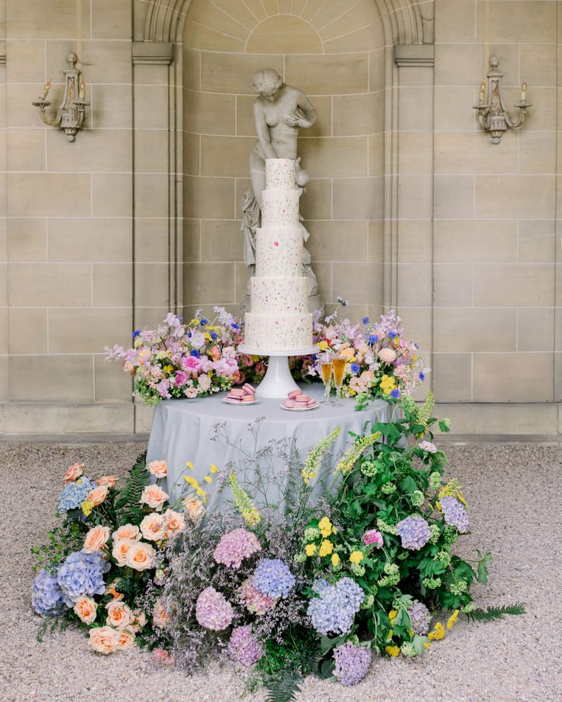 Five-tier wildflower cake on pedestal with lavender hydrangea and peach rose arrangements before stone niche
