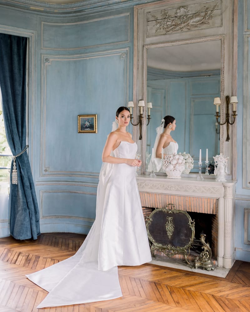 Bride in strapless satin gown with cathedral train standing before marble fireplace in blue paneled chateau room