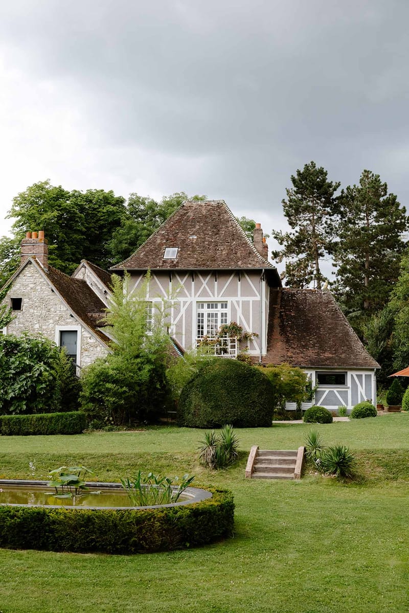 Half-timbered Norman manor house with brick chimneys, small pond, wooden bridge and topiary plantings on manicured grounds