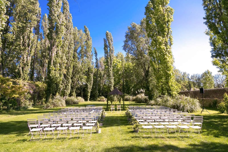 An outdoor ceremony setup photographed in a wide shot on a large lawn enclosed by a stone wall, with tall poplar trees forming a natural backdrop. Rows of white folding chairs are arranged in two sections divided by a central aisle, accommodating approximately 80–100 guests. At the far end of the aisle, a small dark brown canopy or pergola structure serves as the ceremony focal point, flanked by two yellow chairs, likely for the couple. Small potted plants in white pots mark the ends of the aisle rows, and a PA speaker on a stand is visible to the right. The overall decor style is simple and understated, with a neutral white chair palette and minimal floral decoration. Potential venue feature image.