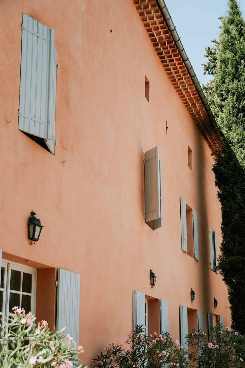 Provencal mas facade with terracotta-orange walls, grey-blue shutters, wrought-iron lanterns, and pink oleander