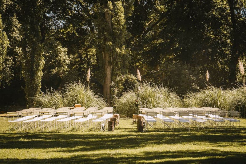Boho ceremony setup with pampas grass clusters, tree-stump aisle markers, and white folding chairs on lawn