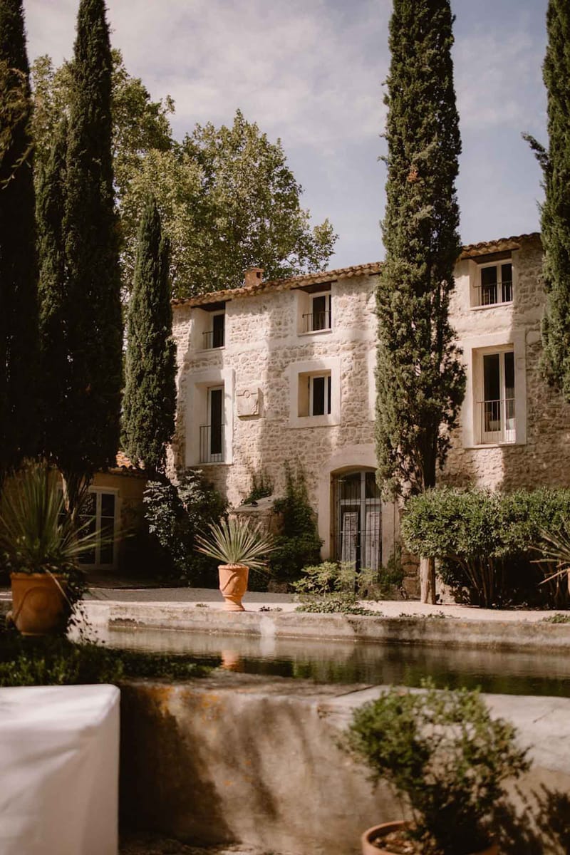 A wide exterior shot of a traditional French stone mas or bastide, shot from the courtyard level. The three-storey building features exposed limestone masonry, terracotta roof tiles, arched doorways with wrought-iron detailing, and small wrought-iron balconies. Tall Italian cypress trees frame the facade on both sides. In the foreground, a long rectangular stone reflecting pool or basin is partially visible, along with terracotta planted pots containing agave-style plants. A white linen-covered surface — likely a cocktail table or furniture — is partially visible at the bottom left, suggesting the space is being set up for a wedding event. The image is a medium-wide architectural shot taken in natural daylight. Potential venue feature image.