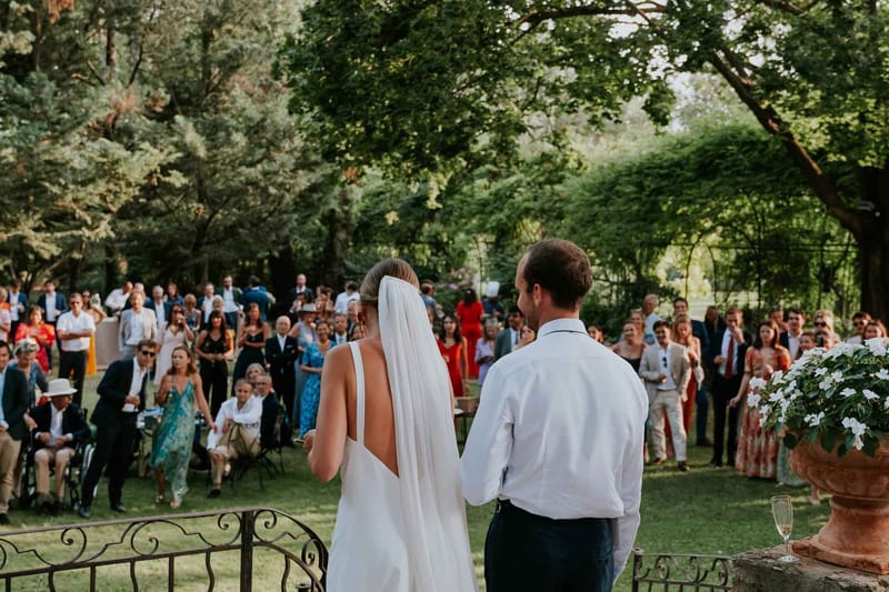 Couple facing 80 guests on garden lawn from elevated terrace with wrought-iron railings
