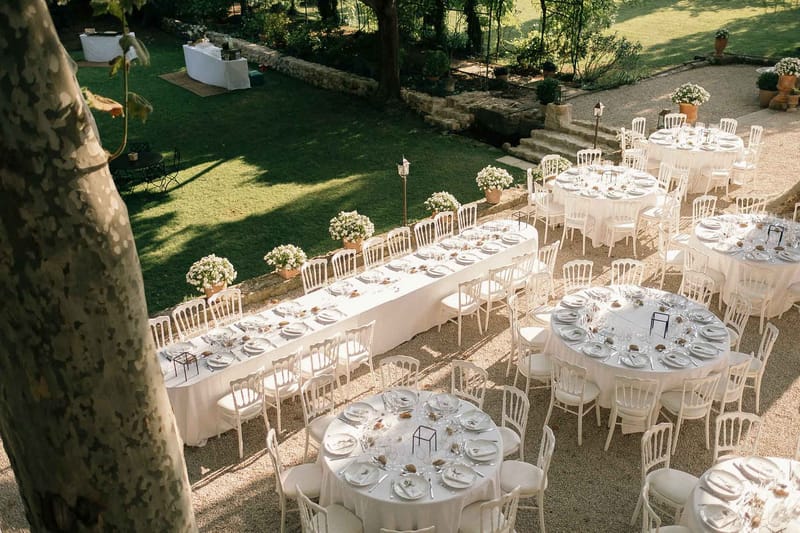 An outdoor wedding reception setup photographed from an elevated angle, showing a gravel courtyard adjacent to a manicured lawn garden. The table arrangement combines one long rectangular table with white linen and several round tables, all dressed in white tablecloths and surrounded by white Napoléon/Chiavari chairs. Each place setting includes white plates, multiple glasses, and small decorative elements including what appear to be geometric black wire cube centerpieces and small natural-toned accents. White flowering plants in terracotta pots are arranged along the low stone border separating the gravel area from the lawn, adding a consistent floral border to the space. A secondary white-draped table is visible further back on the lawn. The overall decor palette is white and neutral with minimal color accents, giving the setup a clean, classic feel. Potential venue feature image.