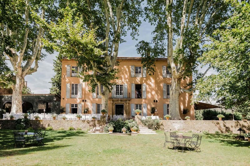 Wide exterior shot of a three-storey Provençal bastide with warm ochre-yellow rendered walls and grey-blue shutters, set up for a wedding reception. The terrace to the left of the building is laid with multiple round tables dressed in white linens and white chairs, arranged under the shade of the building's facade and an adjacent arched stone structure. Terracotta pots with greenery and flowering plants line the front steps and terrace perimeter. Two figures are visible on the first-floor balcony above the main entrance. In the foreground lawn, wrought-iron bistro tables and chairs are placed casually on either side. A canopy or marquee structure is partially visible to the right. The overall decor palette is classic French country with white table linens, terracotta planters, and minimal floral styling. Potential venue feature image.