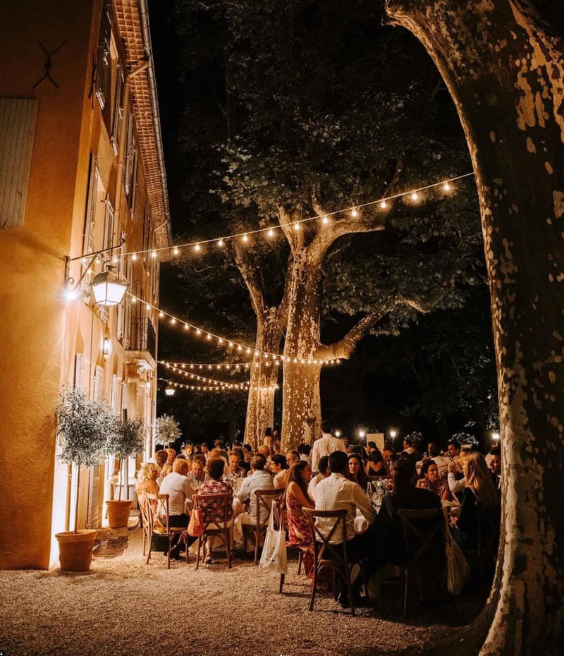 Forty guests dine at long tables under globe lights between plane trees and ochre bastide facade at night