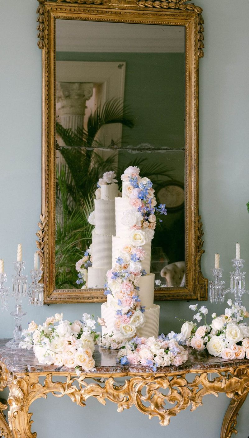Five-tier ribbed buttercream wedding cake with cascading pink roses and blue delphinium on a gilt console table