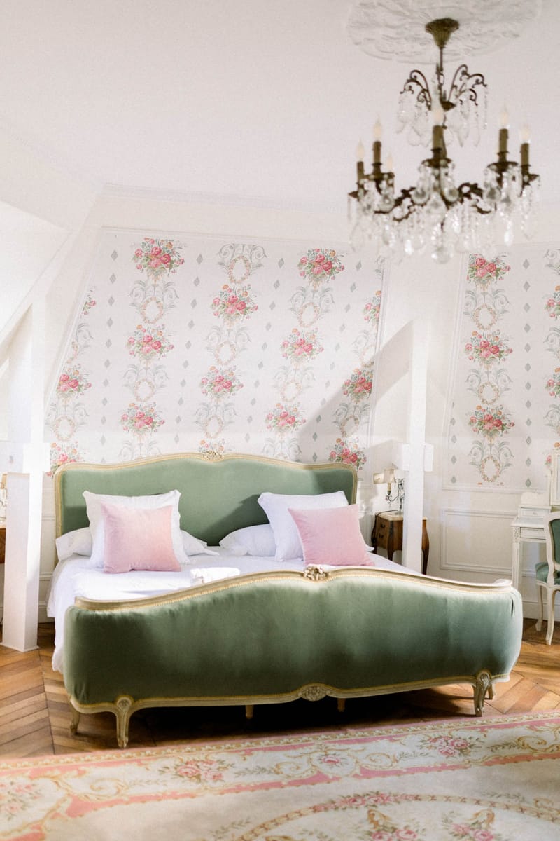 An interior shot of a bedroom suite, likely used as a bridal getting-ready space or accommodation within a French château venue. The room features a Louis XV-style upholstered bed in sage green velvet with a gilt carved wood frame, dressed with white linens and blush pink velvet cushions. The walls are covered in a pink and green floral bouquet wallpaper with a classic French toile-style repeat pattern, and the room is lit by a bronze and crystal chandelier hanging from an ornate plaster ceiling rose. The herringbone parquet wood floor is partially covered by a cream and pink floral Aubusson-style rug, and a green velvet accent chair is visible in the right corner. Wide portrait-orientation shot showing the full bed and room context. Potential venue feature image.
