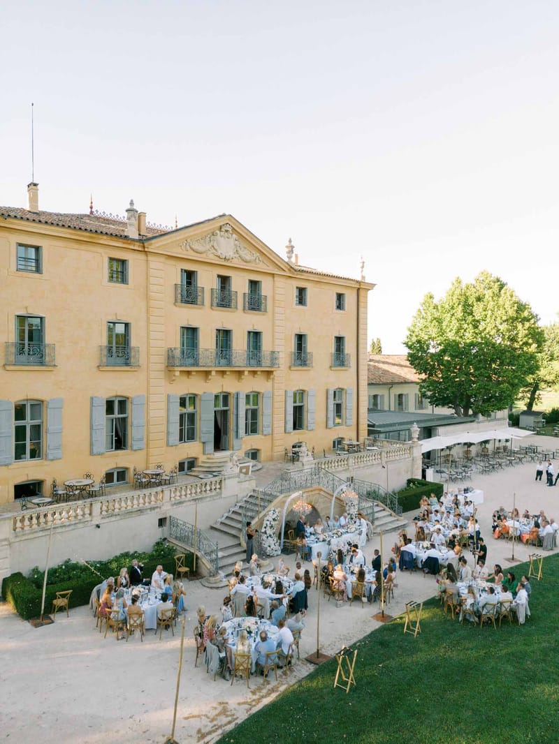 Aerial view of a grand French château with guests seated at round tables across a courtyard and lawn during a reception.