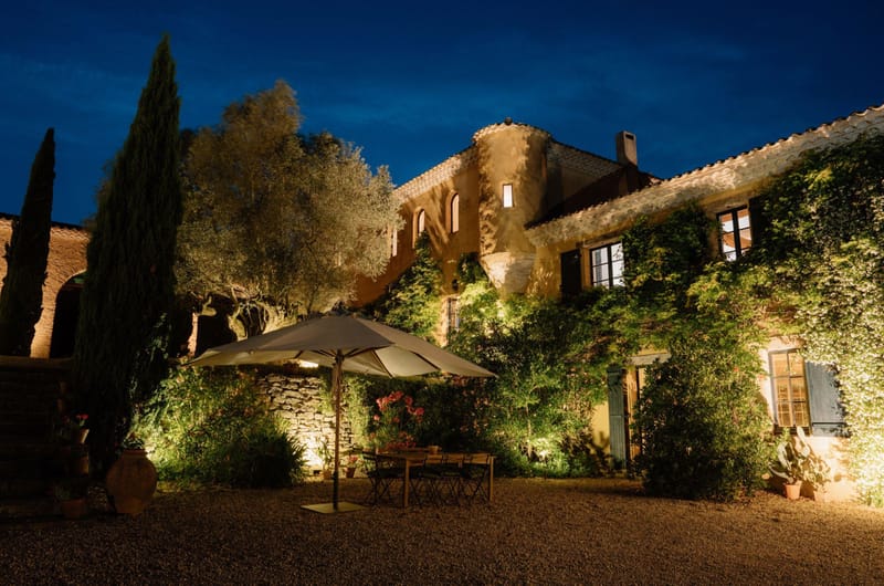 Provencal stone bastide with ochre walls and blue shutters lit by warm uplighting at dusk with gravel courtyard