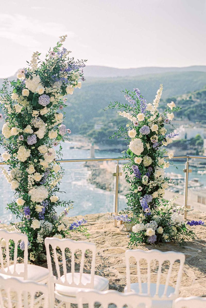 Outdoor wedding ceremony setup on clifftop overlooking water and mountains with floral arch and white chairs