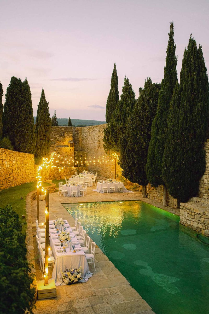 Outdoor reception dinner setup around illuminated pool at stone courtyard venue with cypress trees