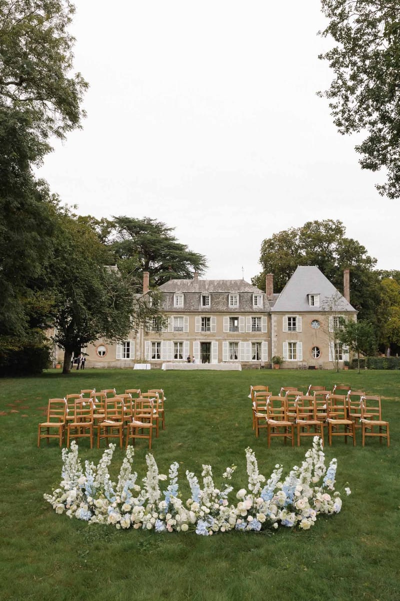 Outdoor wedding ceremony setup on château lawn with floral installation and neoclassical French mansion backdrop