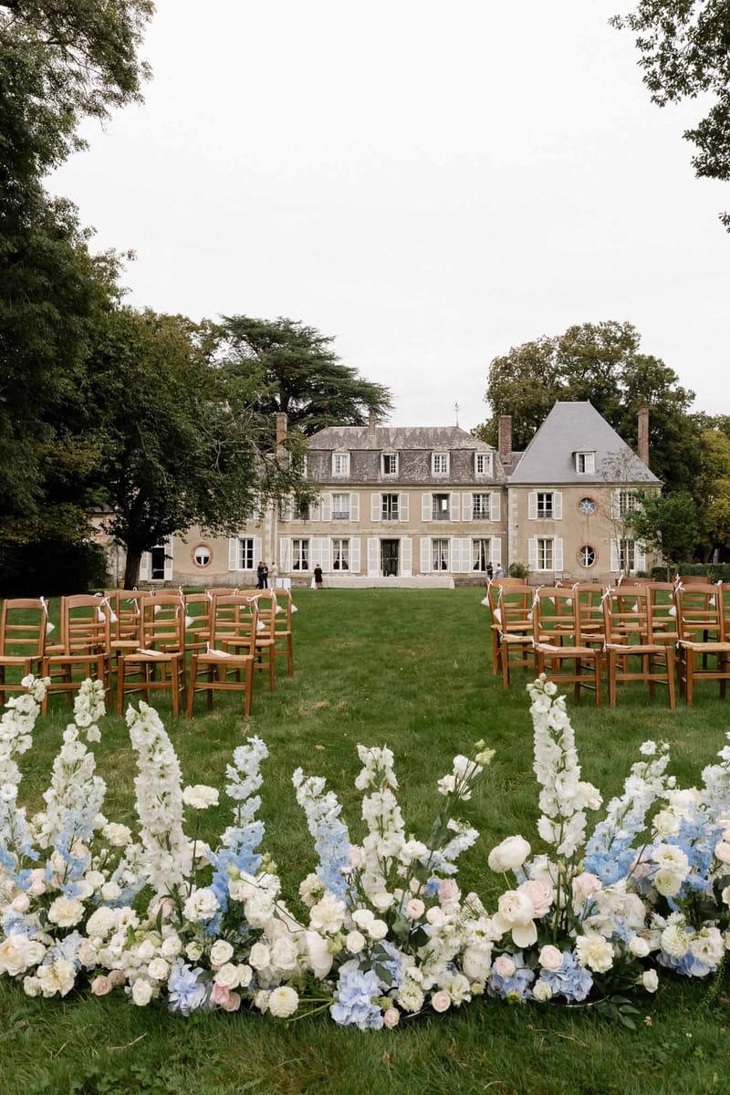 Outdoor wedding ceremony setup on lawn of French château with floral aisle borders and cross-back chairs