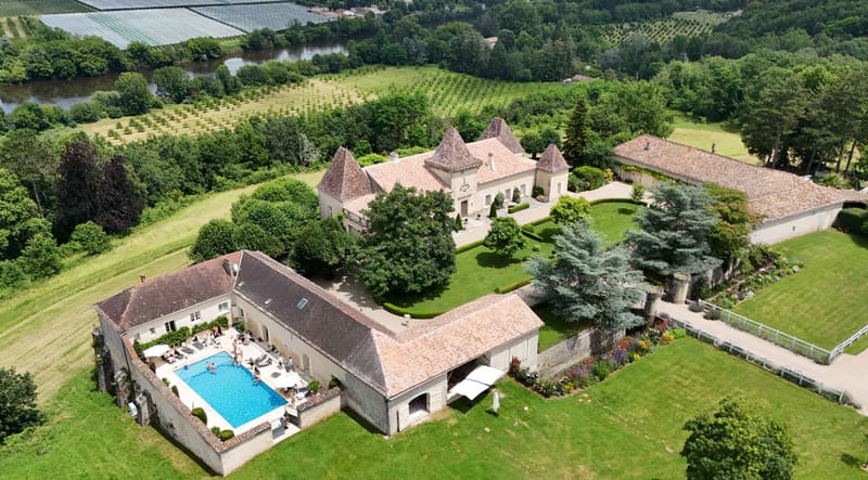 Aerial chateau estate with three conical turrets, pool courtyard, white parasols, and surrounding farmland