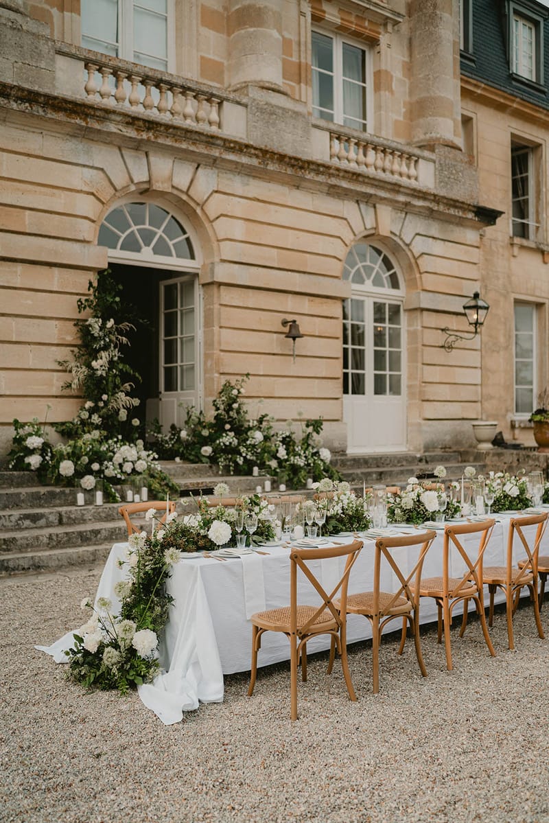 Cross-back chairs at white-clothed table with hydrangea and allium runner before limestone chateau steps