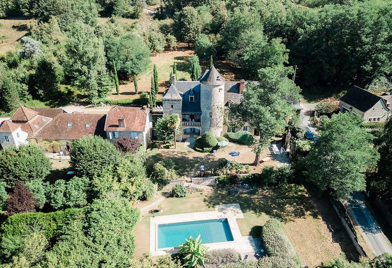 Aerial stone chateau with round tower pool outbuildings cypress trees and dense woodland surroundings
