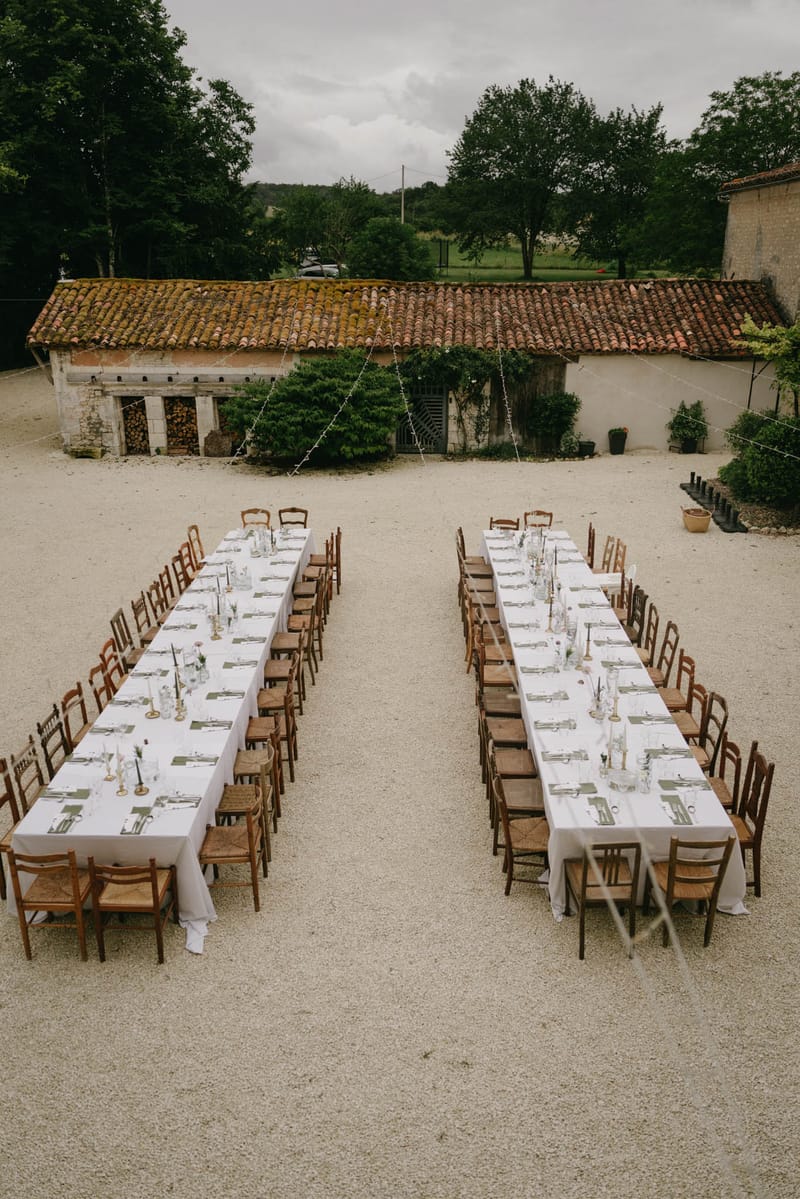 Outdoor reception with two long tables on gravel courtyard, fairy lights, vintage chairs, and stone outbuildings