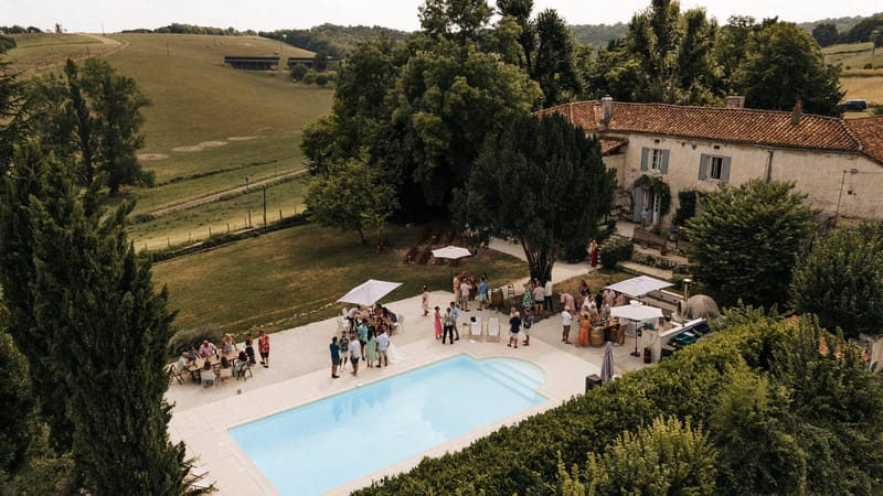 Aerial cocktail hour around pool at stone farmhouse with white umbrellas barrel bar and forty guests in summer attire