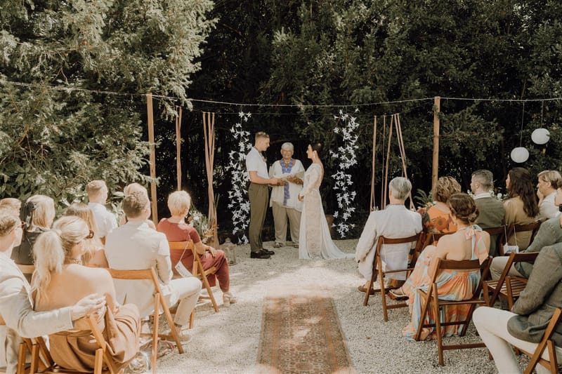 Outdoor boho ceremony with couple at altar beneath fabric streamers and white floral backdrop with seated guests