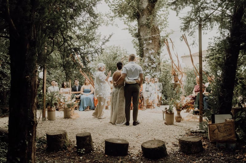 Rustic woodland ceremony with terracotta ribbon streamers, ceramic jug florals, and 20 guests on wooden chairs