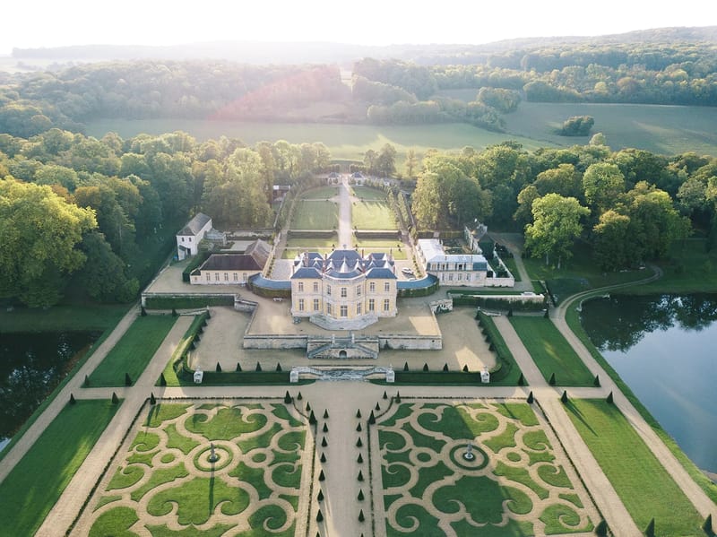Aerial view of yellow stone chateau with mansard roof parterre broderie gardens and tree-lined allee