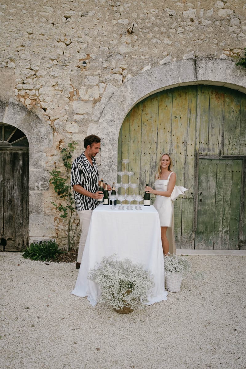 Couple preparing champagne tower on gravel courtyard with baby's breath arrangements beside stone building