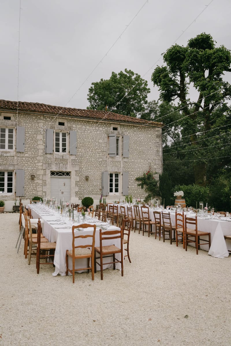 Two long banquet tables in V-shape on gravel courtyard of stone manor with fairy lights and rustic wooden chairs
