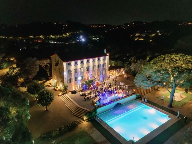 Aerial night reception eighty guests at round tables on terrace with blue pool string lights and purple facade uplighting