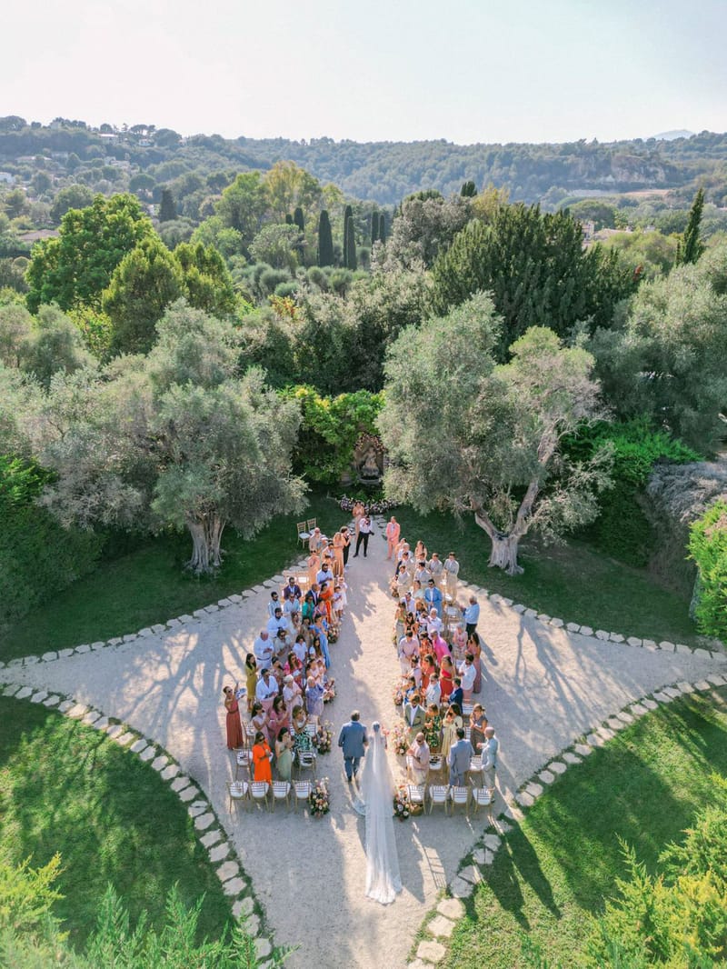 Aerial ceremony on olive tree allee with gold chiavari chairs, warm-toned aisle florals, and rolling hills beyond